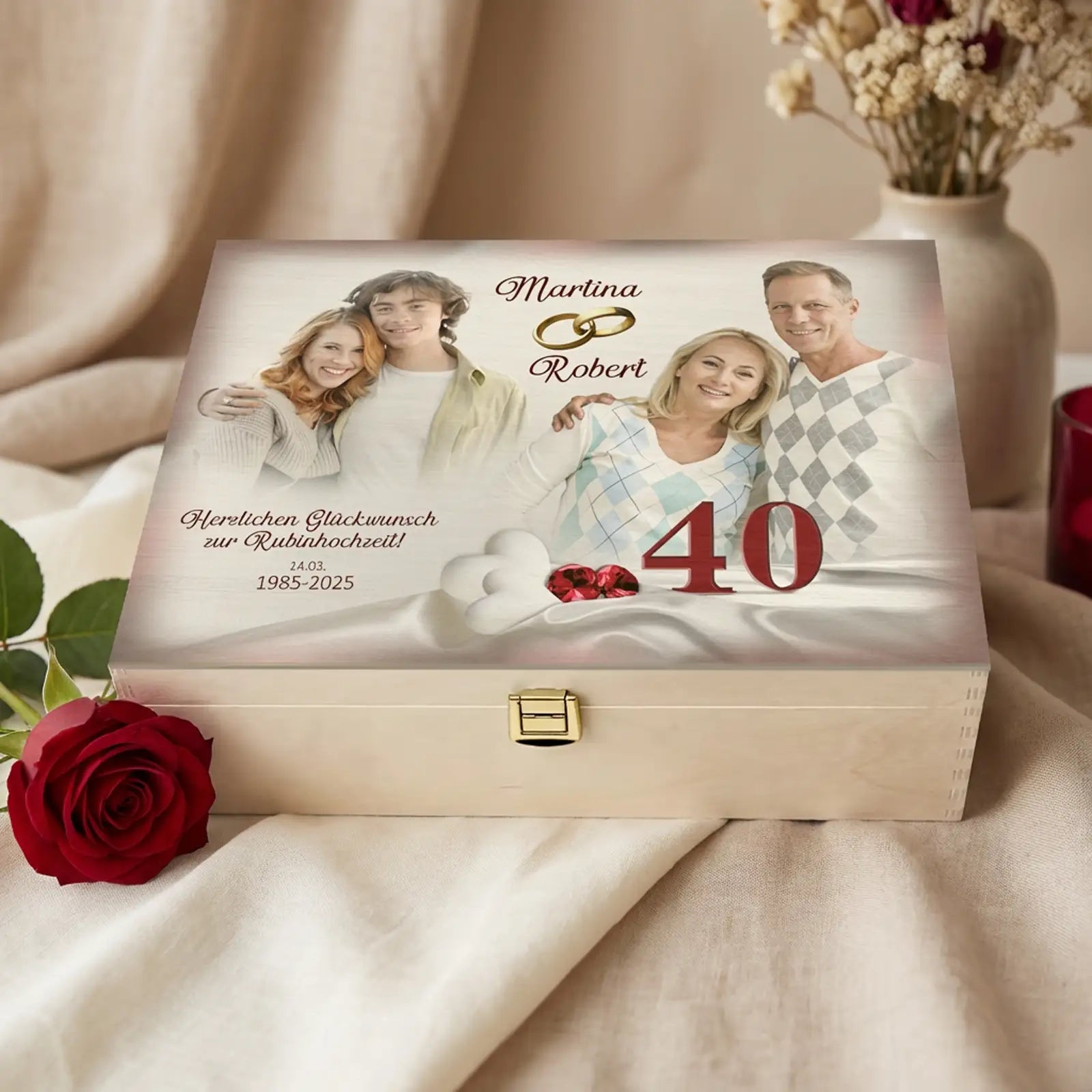 Personalisierte helle Holzbox zur Rubinhochzeit (40 Jahre) mit Fotodruck auf dem Deckel. Auf der Box sind zwei Paarfotos eines Ehepaares zu sehen, dazu die Namen Martina und Robert, goldene Ringe, die Jubiläumszahl 40 sowie ein Glückwunschtext mit Datum. Die Holzbox steht auf einem Stoffuntergrund, daneben liegt eine rote Rose, im Hintergrund sind dezente Wohnaccessoires unscharf zu erkennen. Geschenkidee zum 40. Hochzeitstag.