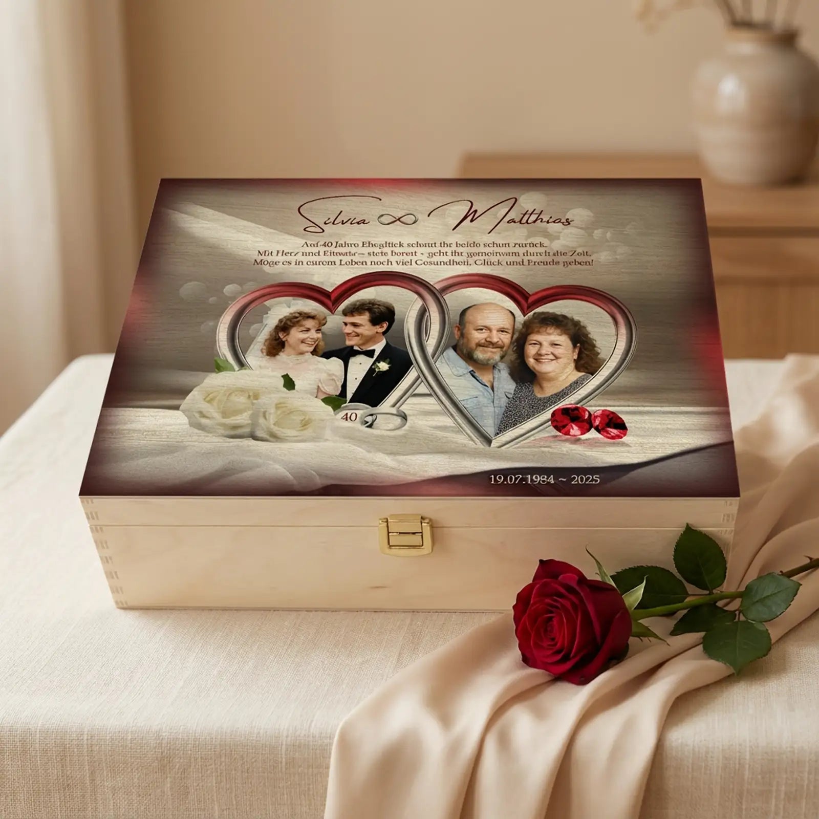 Personalisierte helle Holzbox zur Rubinhochzeit (40 Jahre) mit Fotodruck auf dem Deckel. Auf der Box sind zwei Paarfotos eines Ehepaares in herzförmigen Rahmen zu sehen, dazu die Namen Silvia und Matthias, ein Glückwunschtext, das Hochzeitsdatum 19.07.1984–2025 sowie rote Schmucksteine als Rubin-Symbol. Die Holzbox steht auf einem hellen Stoffuntergrund, rechts davor liegt eine einzelne rote Rose, der Hintergrund ist ruhig und unscharf. Geschenk zum 40. Hochzeitstag.