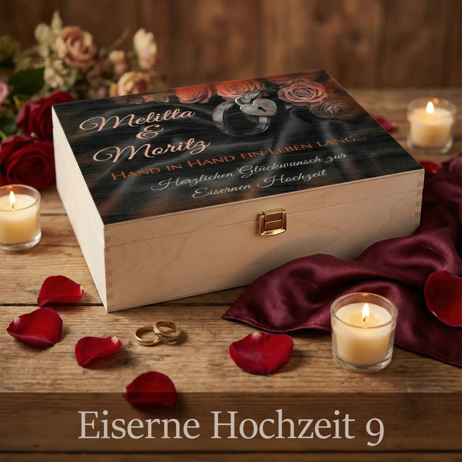Personalisierte Holzbox zur Eisernen Hochzeit mit hellem Holz und Metallverschluss. Auf dem Deckel dunkles, elegantes Design mit Rosen, Herzmotiv und den Namen Melitta und Moritz sowie dem Schriftzug „Hand in Hand ein Leben lang“. Die Box liegt auf einem Holztisch, umgeben von Kerzen, roten Rosenblättern, Stofftuch und Eheringen in stimmungsvollem, romantischem Ambiente als Geschenkidee zum 65. Hochzeitstag.
