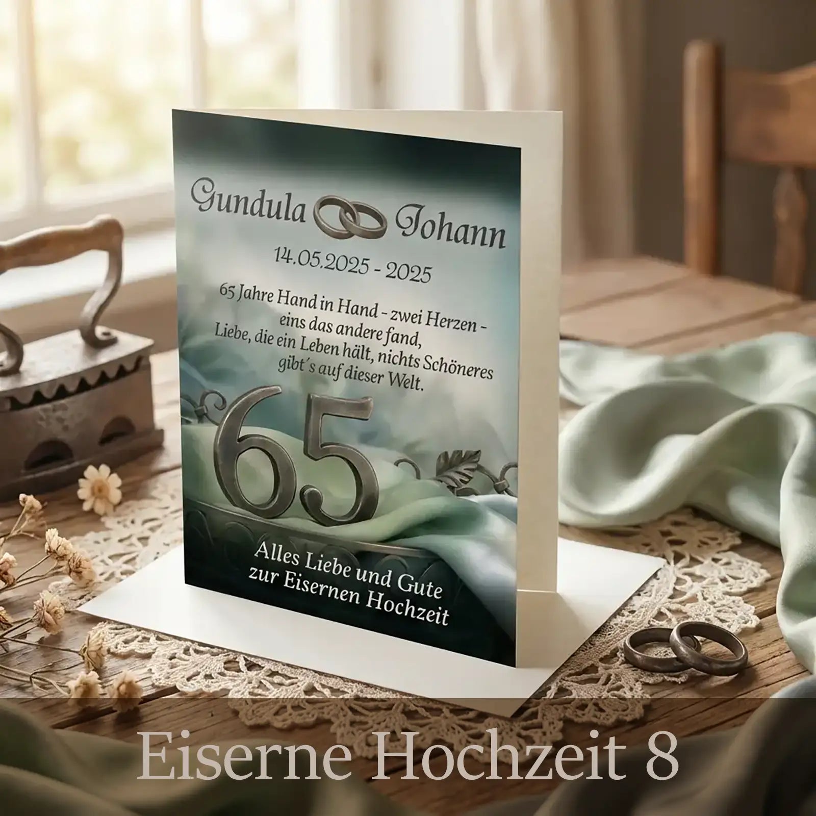 Aufgestellte, personalisierte Glückwunschkarte zur Eisernen Hochzeit auf Holztisch: elegantes Motiv in sanften Grün- und Grautönen mit Stoffstruktur, großer Zahl „65“, ineinander verschlungenen Eheringen und Blätterranken. Vorderseite mit den Namen „Gundula & Johann“, Jubiläumsdatum und einem poetischen Text über 65 Jahre Hand in Hand. Die Karte steht offen auf Spitzenläufer, daneben liegen Trauringe, getrocknete Blumen und ein antikes Bügeleisen im Hintergrund.