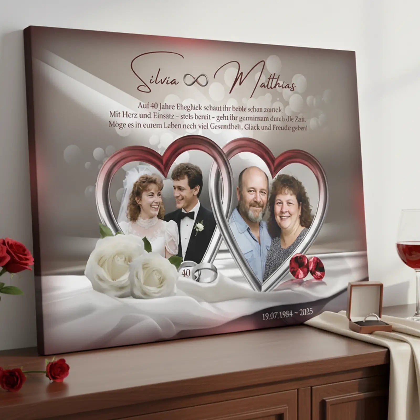 Personalisierte Fotoleinwand zur Rubinhochzeit (40 Jahre) mit zwei Fotos eines Ehepaares in herzförmigen Rahmen: links das Hochzeitsfoto aus jungen Jahren, rechts das Paar im heutigen Alter. Auf der Leinwand stehen die Namen Silvia und Matthias, ein Glückwunschtext zum 40. Hochzeitstag sowie das Datum 19.07.1985–2025. Dekorative Elemente sind weiße Rosen, rote Schmucksteine und ein heller Stoffhintergrund. Die Leinwand steht auf einem Sideboard